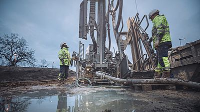 Baustelle in der Dämmerung. Zwei Arbeiter in Sicherheitskleidung bei der Tiefenbohrung der Wärmesonden im VILLAGE IM DRITTEN.