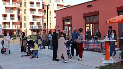 Mehrere Menschen stehen vor einem kleinen Gebäude und unterhalten sich, im Hintergrund ist ein neuer Stadtteil zu sehen.