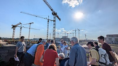 Mehrere Menschen stehen outdoor um einen Tisch mit Planungsunterlagen herum, im Hintergrund sind Baukräne zu sehen.